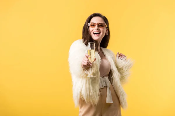 Счастливая тусовщица в искусственной меховой куртке и солнцезащитных очках с шампанским, изолированным на желтом — стоковое фото