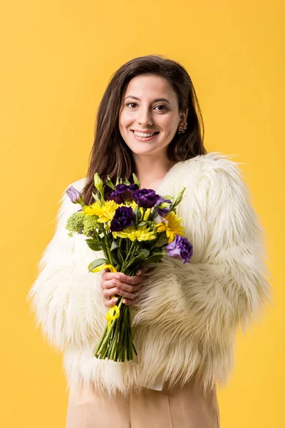 Усміхнена дівчина в штучній шубці тримає букет квітів ізольовано на жовтому — стокове фото