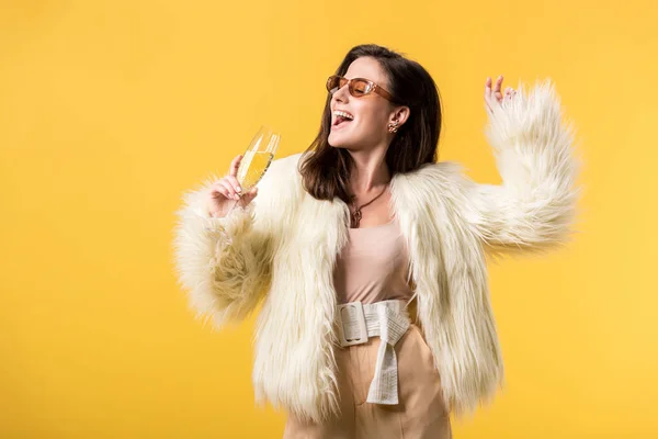 Счастливая тусовщица в искусственном меховом пиджаке и солнцезащитных очках танцующая с шампанским изолированным на желтом — стоковое фото