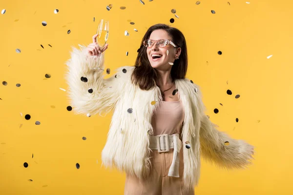 Excitée partie fille en fausse fourrure veste et lunettes de soleil debout sous confettis avec champagne isolé sur jaune — Photo de stock