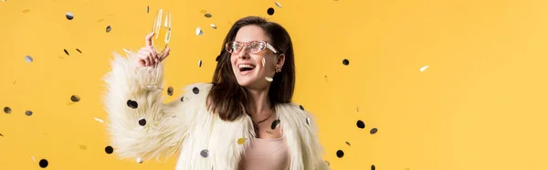 Plano panorámico de chica fiesta excitada en chaqueta de piel sintética y gafas de sol de pie debajo de confeti con champán aislado en amarillo - foto de stock
