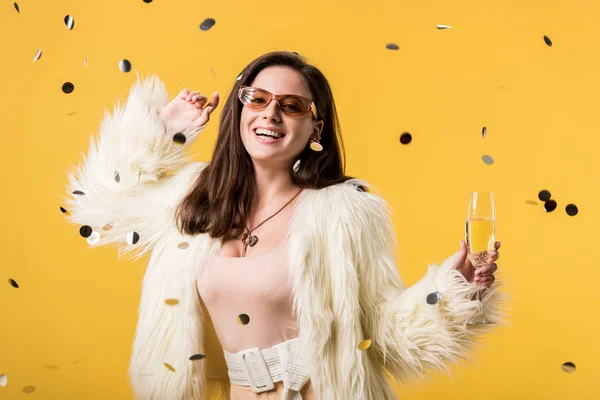 Ragazza partito eccitato in giacca di pelliccia sintetica e occhiali da sole in piedi sotto coriandoli con champagne isolato su giallo — Foto stock