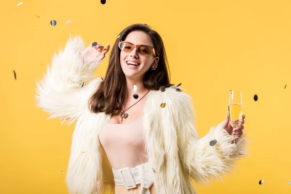 Excited party girl in faux fur jacket and sunglasses standing under confetti with champagne isolated on yellow — Stock Photo
