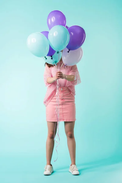 Chica en traje rosa sosteniendo globos en frente de la cara sobre fondo turquesa - foto de stock