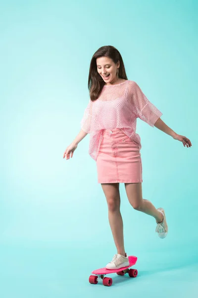 Happy girl in pink outfit riding penny board on turquoise background — Stock Photo