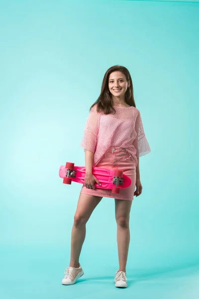 Happy girl in pink outfit holding penny board on turquoise background — Stock Photo