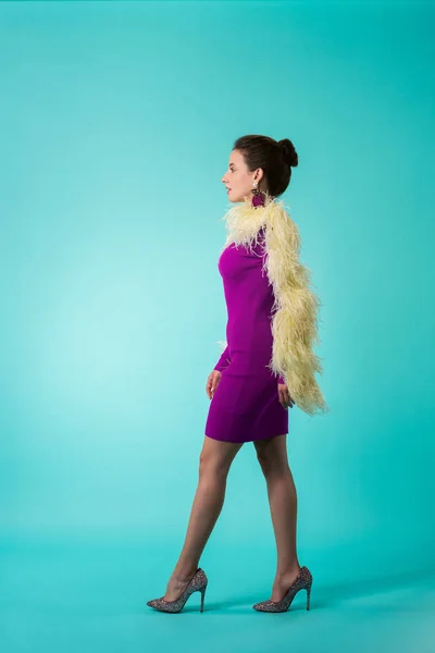Side view of party girl in purple dress with feathers walking on turquoise background — Stock Photo
