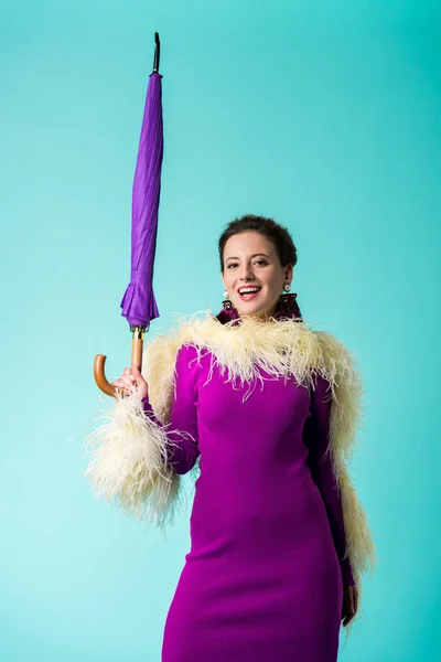 Chica fiesta feliz en vestido púrpura con plumas sosteniendo paraguas aislado en turquesa - foto de stock