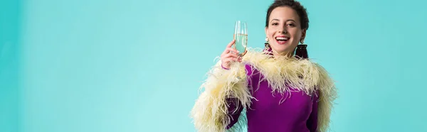 Panoramic shot of happy party girl in purple dress with feathers holding glass of champagne isolated on turquoise — Stock Photo