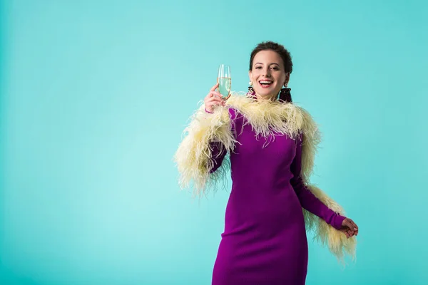 Chica fiesta feliz en vestido púrpura con plumas sosteniendo copa de champán aislado en turquesa — Stock Photo
