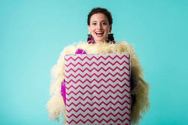 Счастливая тусовщица в фиолетовом платье с перьями, держащих огромный подарочный коробок изолирован на бирюзовый — стоковое фото