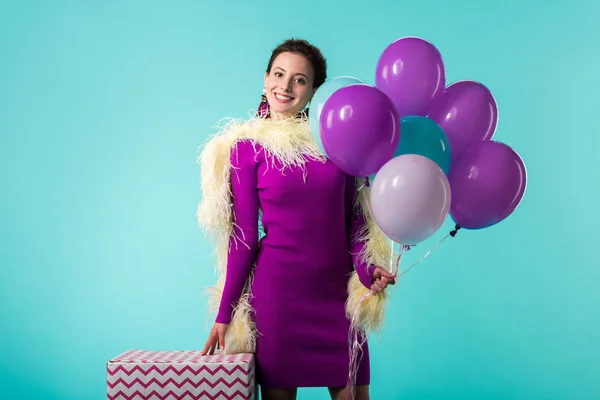 Chica fiesta feliz en vestido púrpura con plumas sosteniendo globos cerca de la caja de regalo enorme aislado en turquesa - foto de stock