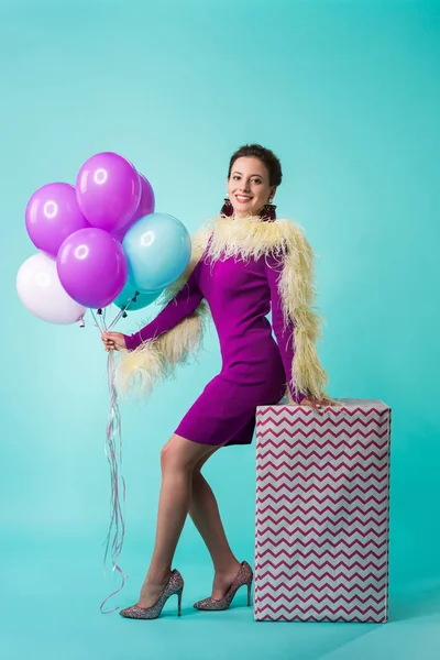 Chica de fiesta feliz en vestido púrpura con plumas sosteniendo globos en la caja de regalo enorme en turquesa - foto de stock