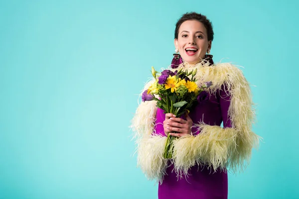 Щаслива вечірка дівчина в фіолетовій сукні з пір'ям, що тримає букет квітів ізольовано на бірюзовому — стокове фото