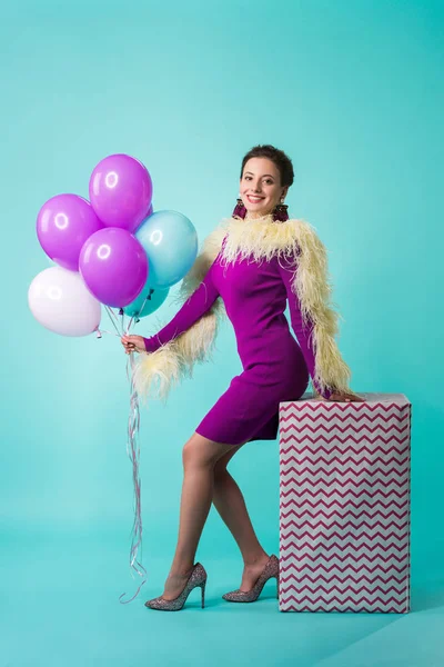 Happy party girl in purple dress with feathers holding balloons on huge gift box on turquoise — Stock Photo