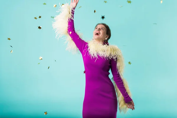 Счастливая тусовщица в фиолетовом платье с перьями танцующими под падающими конфетти изолированы на бирюзовый — стоковое фото
