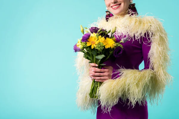Vista ritagliata di ragazza partito sorridente in abito viola con piume che tengono bouquet di fiori isolati su turchese — Foto stock