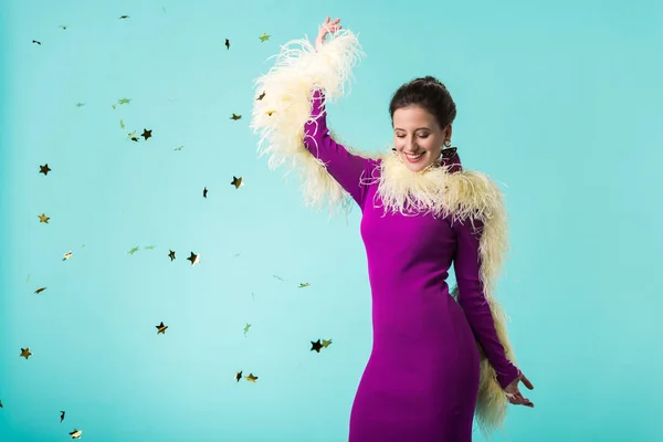 Happy party girl in purple dress with feathers dancing under falling confetti isolated on turquoise — Stock Photo