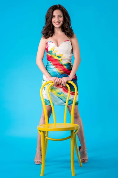 Sonriente elegante mujer joven en vestido posando cerca de la silla amarilla sobre fondo azul - foto de stock