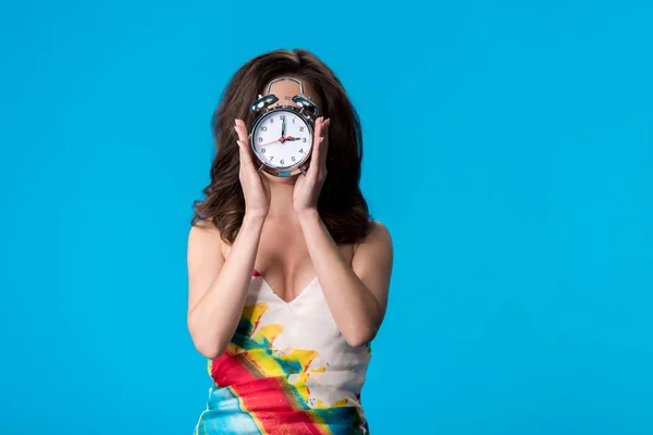 Elegant young woman in dress holding silver alarm clock in front of face isolated on blue — Stock Photo