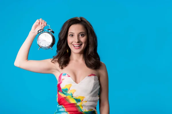 Happy elegant young woman in dress holding silver alarm clock isolated on blue — Stock Photo