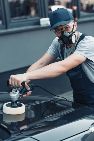 Car cleaner in respirator and protective glasses looking at camera while polishing car with polish machine — Stock Photo