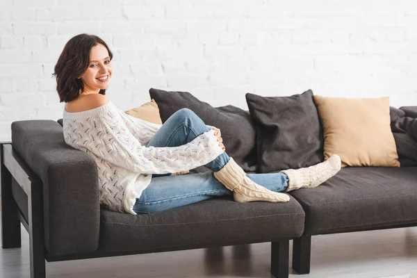 Jolie femme brune assise sur un canapé avec des oreillers dans un salon confortable — Photo de stock