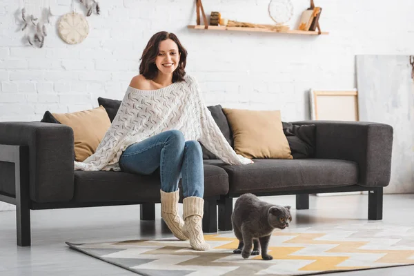 Belle femme heureuse assise sur un canapé avec chat pliant écossais sur le sol — Photo de stock