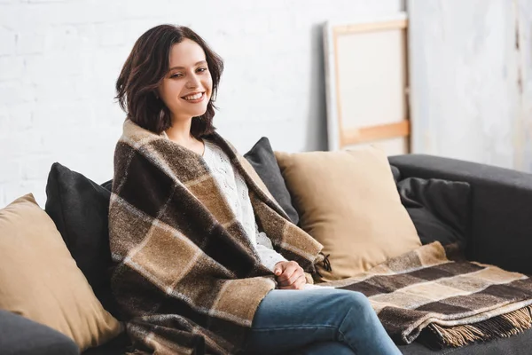 Happy brunette woman in blanket sitting on sofa in cozy living room — Stock Photo