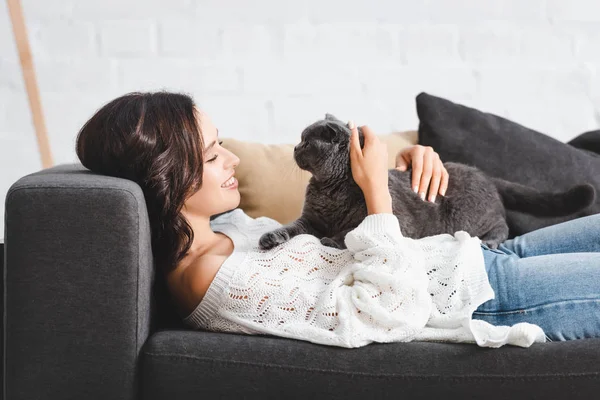 Красивая счастливая брюнетка женщина лежит на диване со шотландской складной кошкой в гостиной — стоковое фото