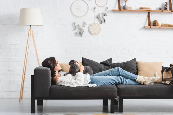 Belle femme couchée sur canapé avec chat pliant écossais dans le salon — Photo de stock