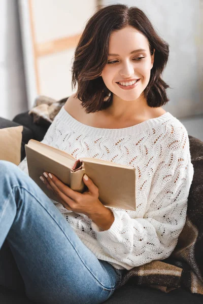 Красивая жизнерадостная девушка, читающая книгу на диване дома — стоковое фото