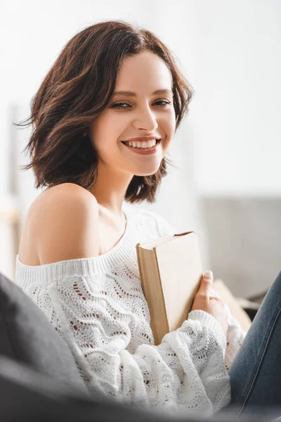 Beau livre de lecture fille souriante sur canapé à la maison — Photo de stock