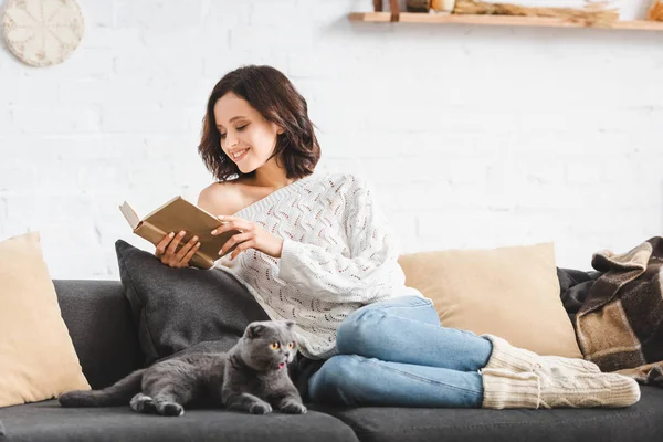 Щаслива жінка читає книгу з шотландським складним котом на дивані — стокове фото