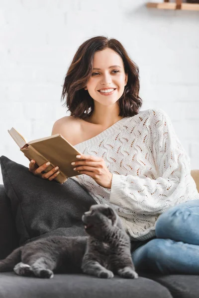 Усміхнена жінка читає книгу з шотландським складним котом на дивані — стокове фото