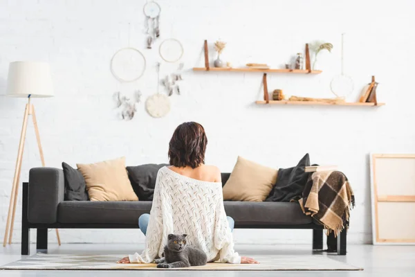 Rückansicht einer Frau, die mit grauer Katze im Wohnzimmer mit Traumfängern auf dem Boden sitzt — Stockfoto