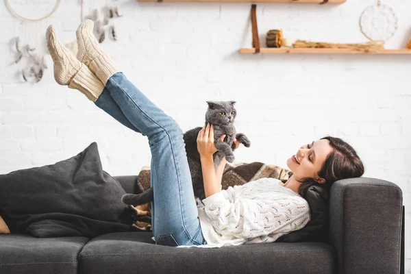 Belle fille gaie couchée sur le canapé avec chat pliant écossais — Photo de stock