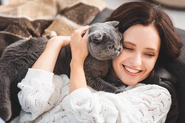 Красивая улыбающаяся девушка лежит на диване со шотландской складной кошкой — стоковое фото