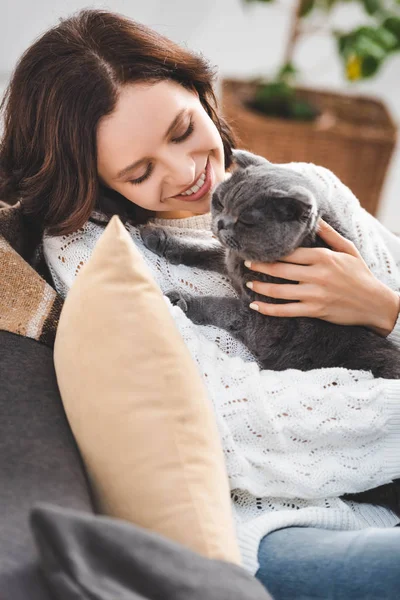 Hermosa joven sentada en sofá con escocés plegable gato - foto de stock