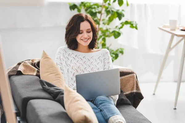 Hermosa chica alegre utilizando el ordenador portátil en el sofá en la acogedora sala de estar - foto de stock