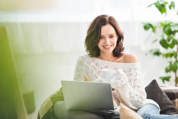 Atractiva chica sonriente con taza de café de usar el ordenador portátil en casa acogedora - foto de stock