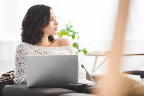 Menina pensativa com copo de bebida quente usando laptop em casa aconchegante — Fotografia de Stock