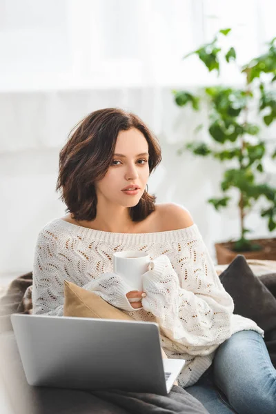 Atractiva chica pensativa con taza de café utilizando el ordenador portátil en casa acogedora - foto de stock