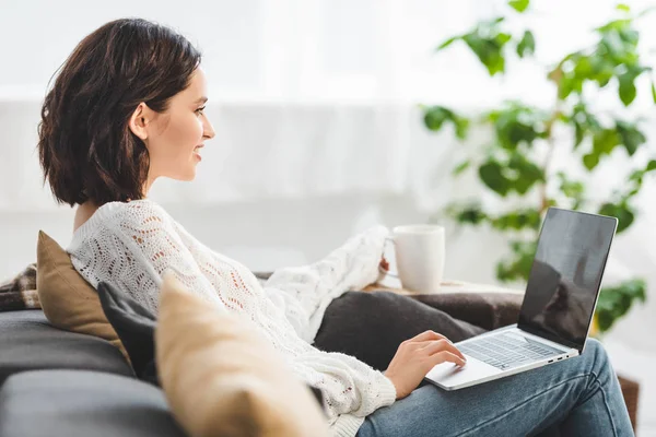 Jolie fille souriante avec une tasse de café en utilisant un ordinateur portable à la maison confortable — Photo de stock