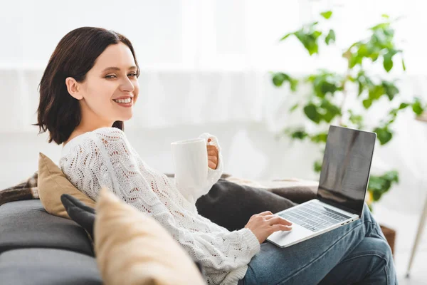 Chica alegre con taza de té usando el ordenador portátil en casa acogedora — Stock Photo