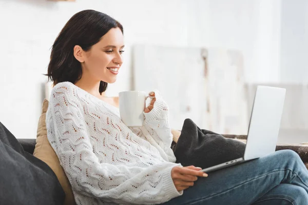 Attraente sorridente ragazza con tazza di caffè guardando film sul computer portatile a casa accogliente — Foto stock