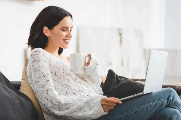 Atractiva chica sonriente con taza de café viendo la película en el ordenador portátil en casa acogedora — Stock Photo