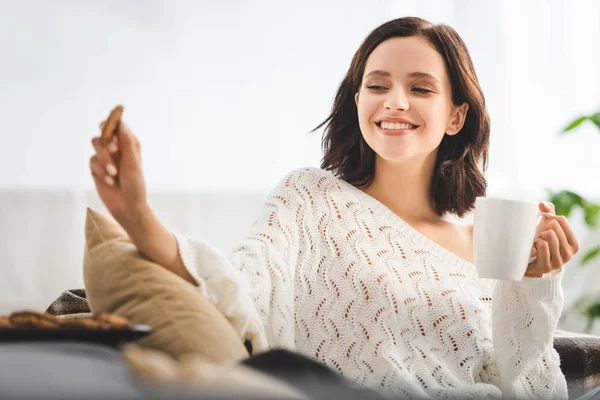 Schöne glückliche Frau sitzt auf Sofa mit Keksen und Kaffeetasse — Stockfoto