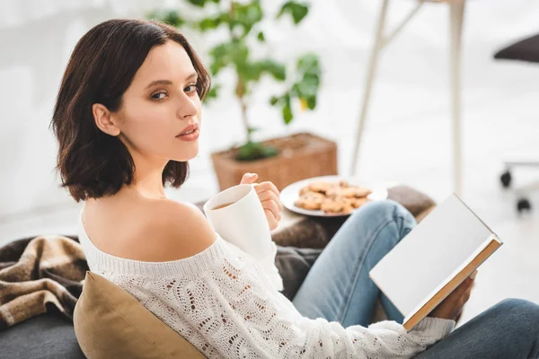 Schönes brünettes Mädchen liest Buch auf Sofa mit Kaffee und Keksen — Stockfoto