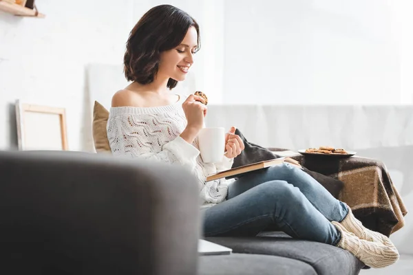Hermosa chica feliz lectura libro en el sofá con café y galletas - foto de stock